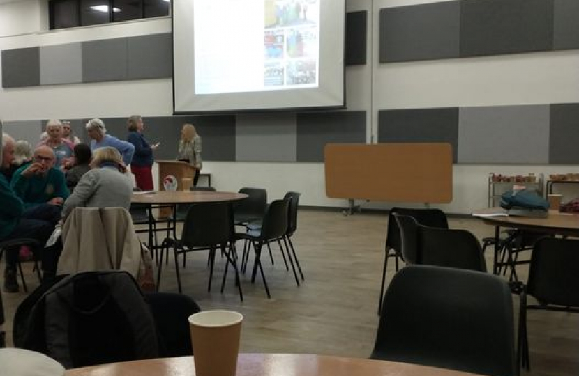 A projector screen with a blurred image, there is a table in the foreground with a paper coffee cup on it and another two tables in the background. One of the tables has a number of people sitting round it.