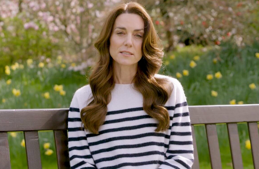 Princess of Wales sitting on a bench. There is grass and flowers behind her and she is wearing a white jumper with dark horizontal stripes.