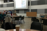 A projector screen with a blurred image, there is a table in the foreground with a paper coffee cup on it and another two tables in the background. One of the tables has a number of people sitting round it.