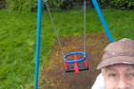 Cllr Tim Nelson selfie in front of refurbished swing. He is wearing a white jacket and beige hat. He is smiling.